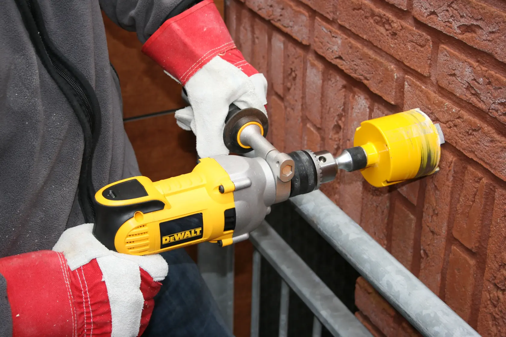 Uma pessoa usando luvas de proteção está utilizando uma furadeira elétrica amarela DEWALT com uma serra copo para perfurar uma parede de tijolos.