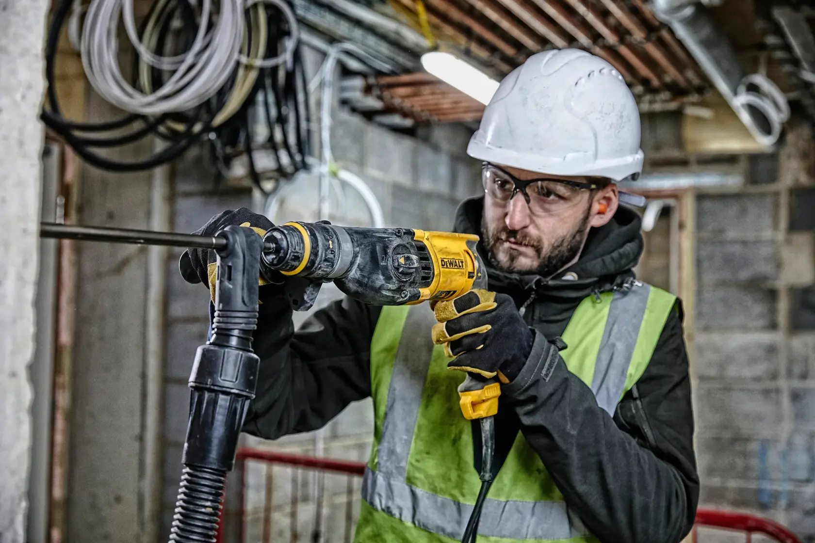 Uma pessoa usando capacete de segurança, luvas e colete refletivo está utilizando um martelete rotativo DEWALT D25134K em um canteiro de obras. Ao fundo, há cabos e tubulações expostas em um prédio inacabado.