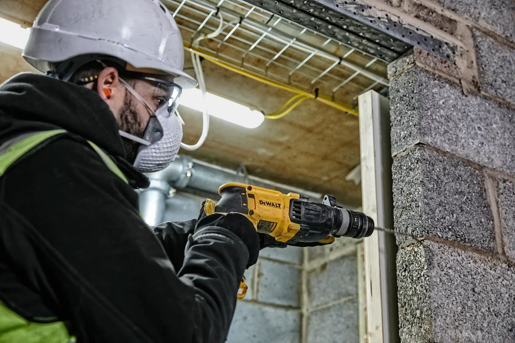 Um trabalhador usando capacete de segurança e luvas utiliza um martelete rotativo DEWALT D25134K para perfurar uma parede de concreto em um canteiro de obras.