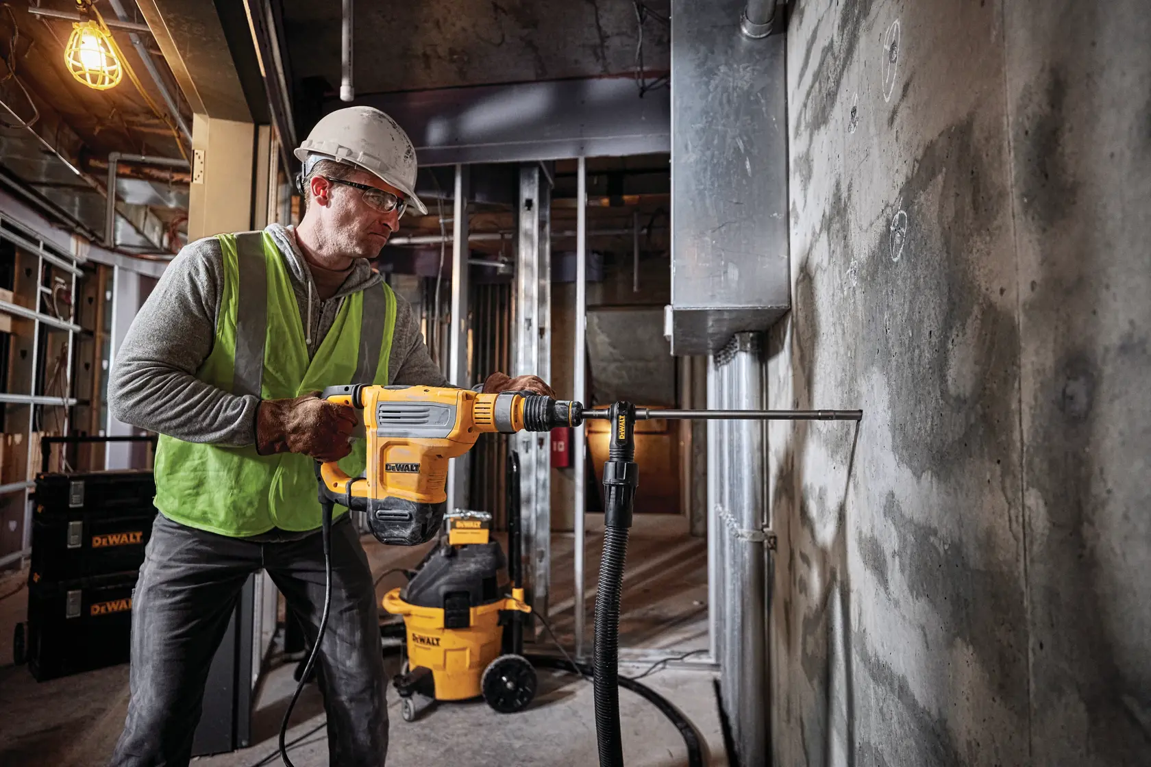 Uma pessoa usando equipamento de segurança e capacete opera um martelo rotativo DEWALT D25614K em uma parede de concreto em um local de construção. Equipamentos e ferramentas DEWALT são visíveis ao fundo.