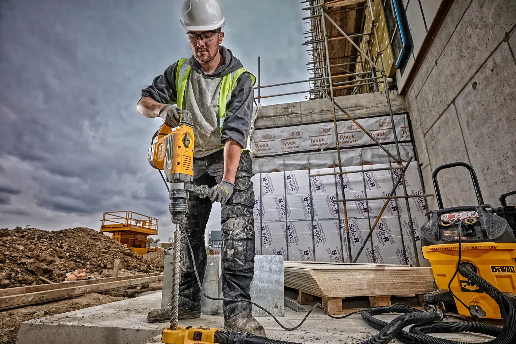 Um trabalhador da construção civil com capacete de segurança e colete de alta visibilidade opera um martelo rotativo DEWALT (SKU: D25733K) em um canteiro de obras. O rosto do trabalhador está desfocado, e há equipamentos de construção, andaimes e materiais isolantes ao fundo.