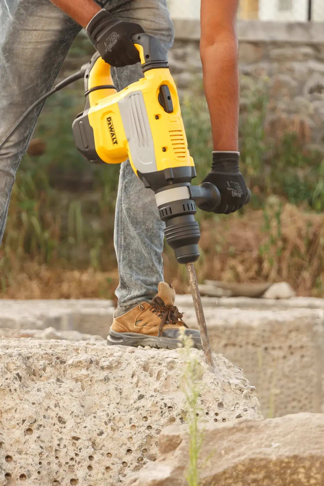 Uma pessoa utiliza um martelete rotativo amarelo DEWALT (modelo D25733K) para quebrar concreto ao ar livre. A pessoa está usando luvas, jeans e botas de trabalho.