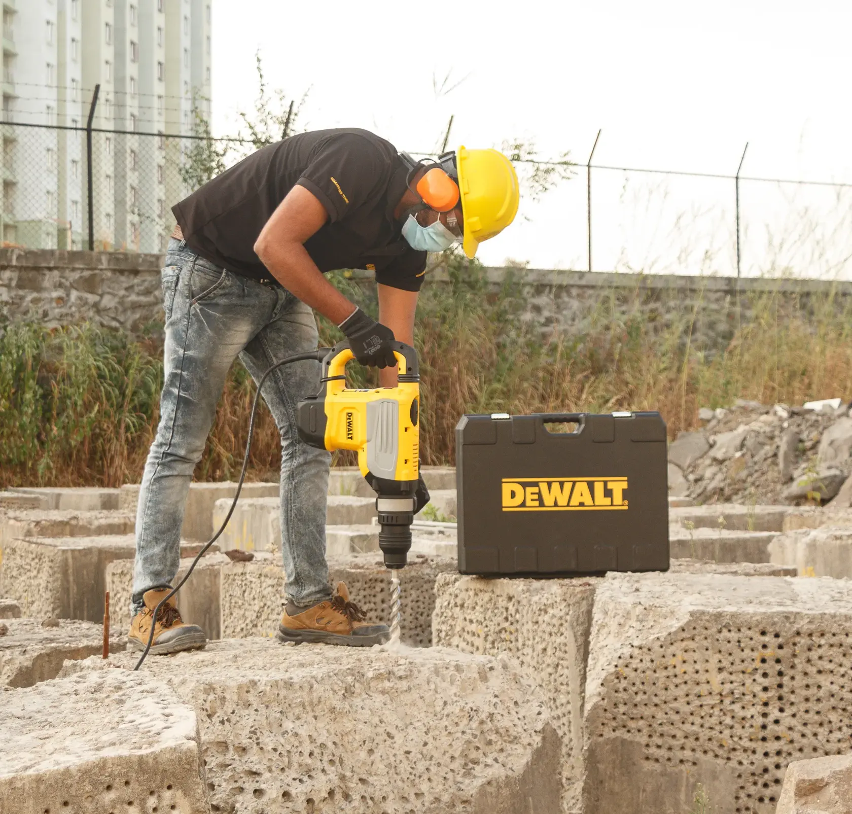 Uma pessoa usando equipamentos de segurança, incluindo capacete, luvas, proteção auricular e máscara, utiliza um martelete DEWALT em um grande bloco de concreto. Uma maleta de ferramentas DEWALT está colocada próxima.