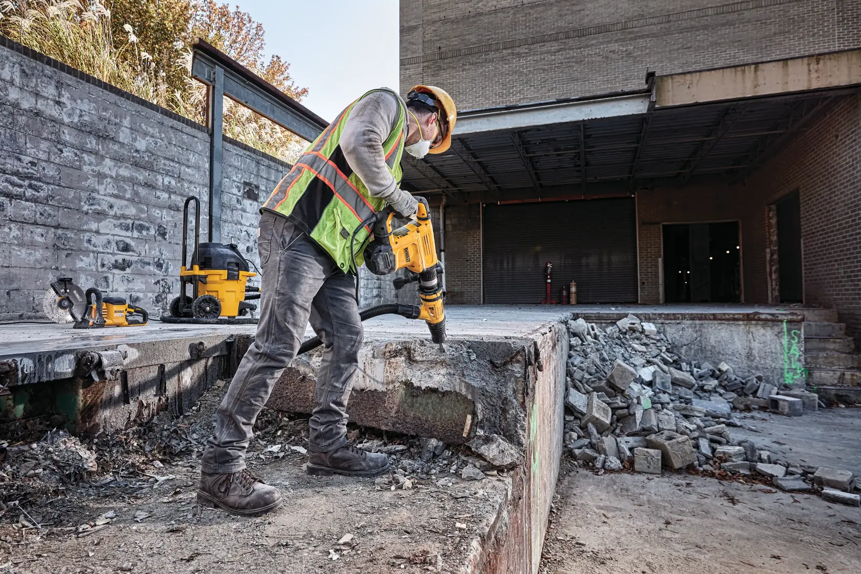 Um trabalhador da construção civil usando equipamentos de segurança utiliza um martelo demolidor DEWALT em uma estrutura de concreto em uma obra. Concreto quebrado e entulhos são visíveis ao redor da área.