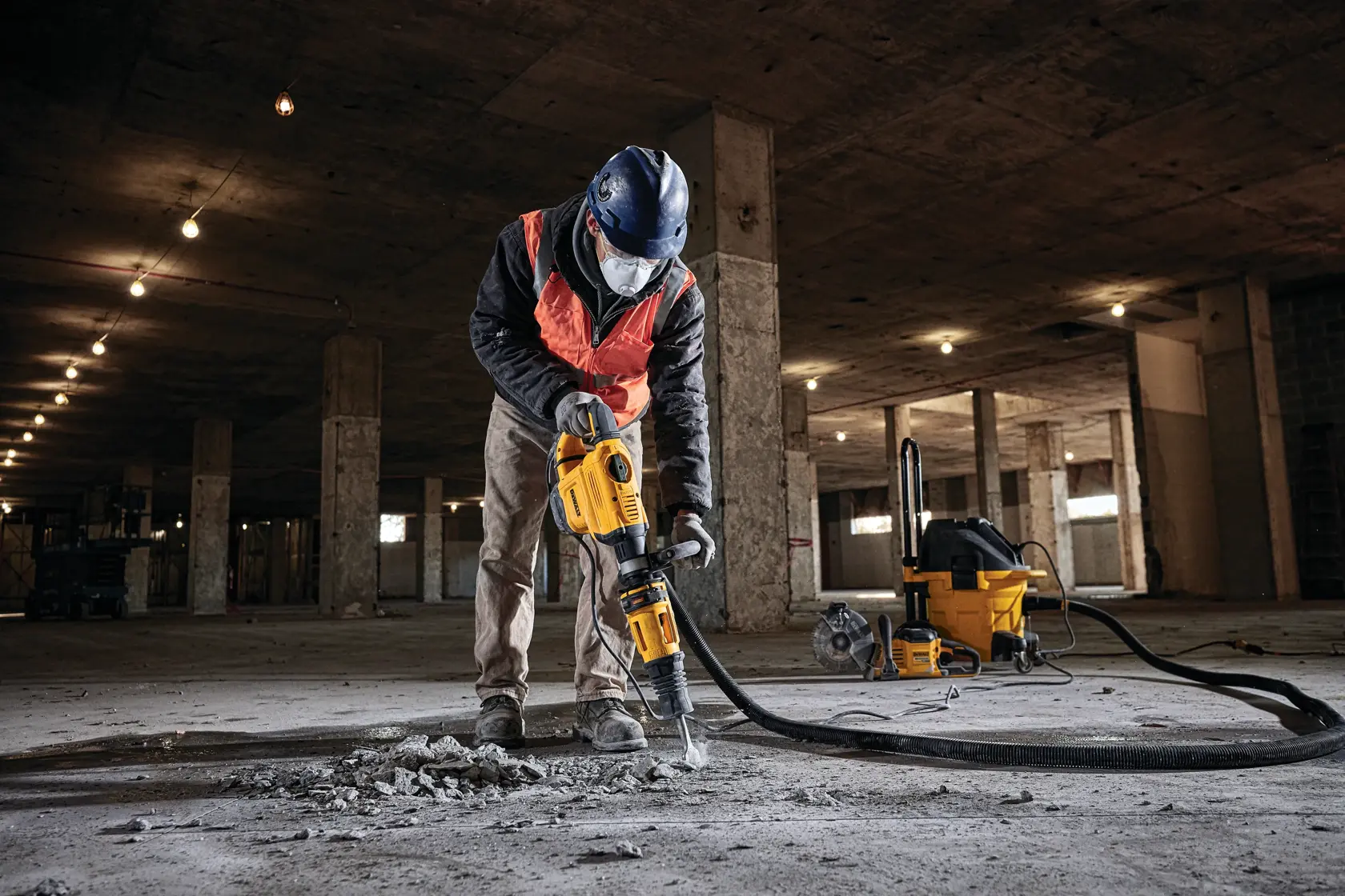 Uma pessoa usando equipamento de segurança, incluindo capacete e máscara facial, utiliza um martelo demolidor amarelo DEWALT para quebrar concreto no chão de um grande canteiro de obras. Uma máquina de aspiração amarela DEWALT é visível ao fundo.
