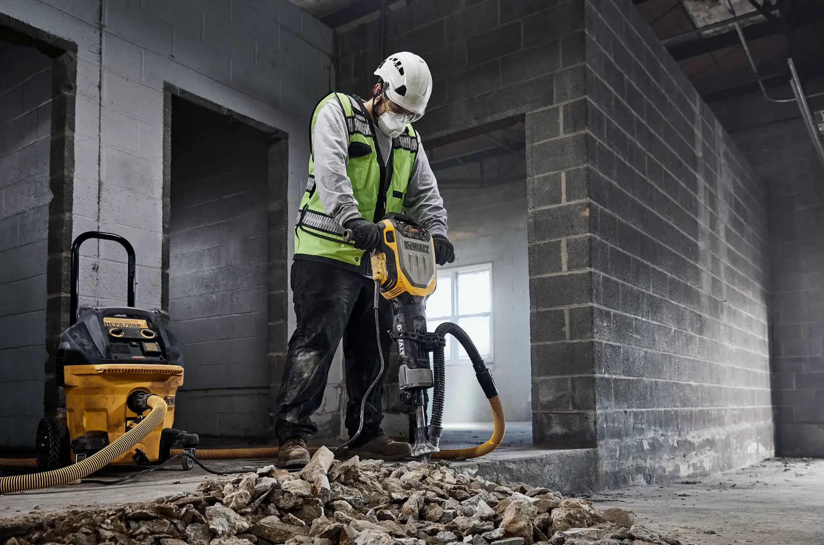 Um trabalhador da construção civil com colete de segurança e capacete está usando um martelete DEWALT (modelo D25966) para quebrar concreto dentro de um prédio inacabado. Um extrator de pó DEWALT está conectado ao martelete, e pedaços de concreto quebrado estão espalhados no chão.