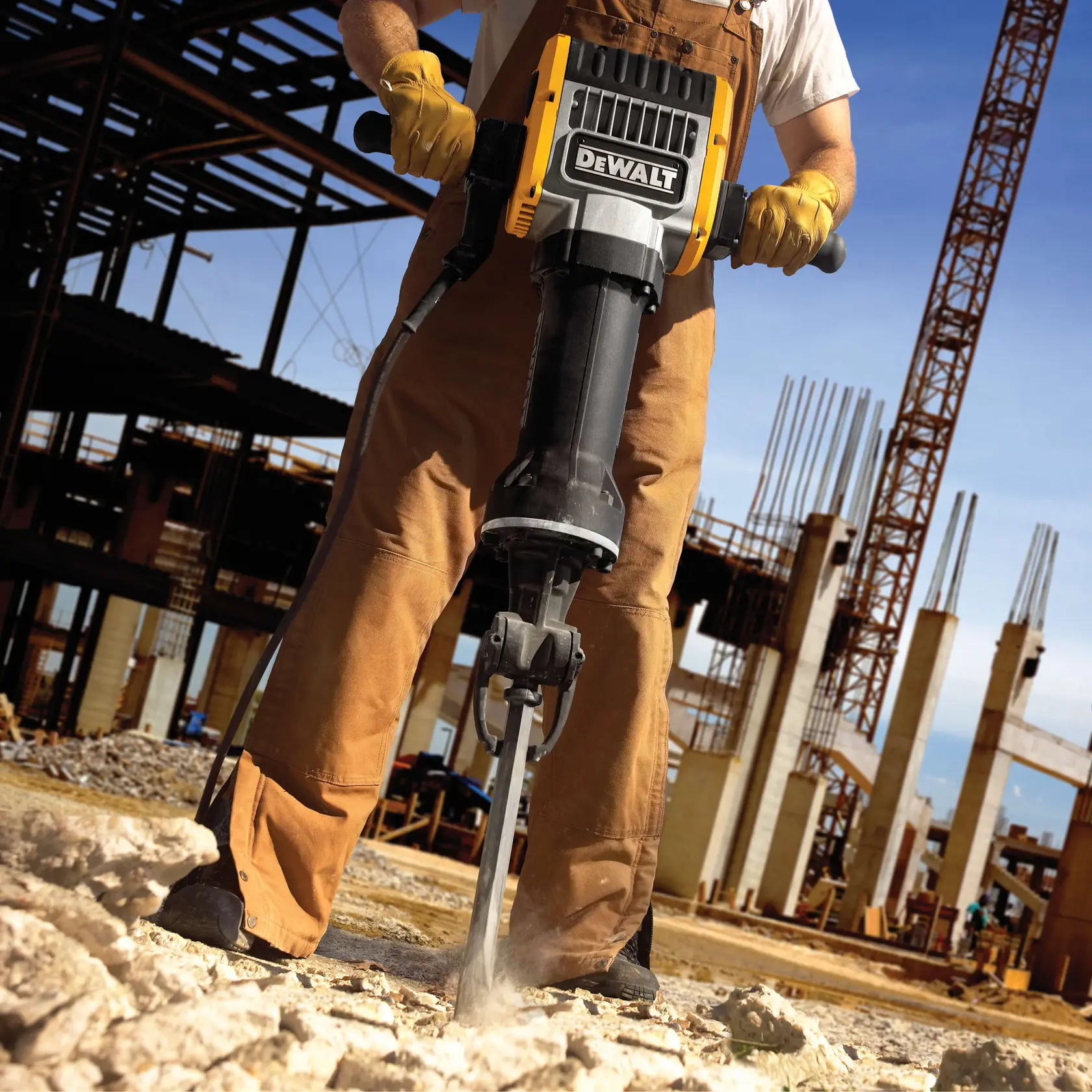 Uma pessoa usando luvas de trabalho e macacão opera um martelete DEWALT, quebrando concreto em um canteiro de obras com estruturas de aço e colunas visíveis ao fundo.