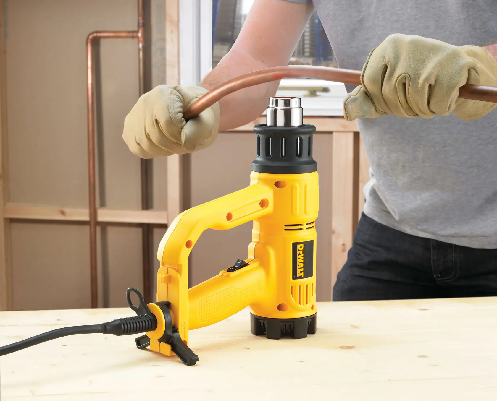 Uma pessoa com luvas usa um soprador térmico DEWALT para dobrar um tubo de cobre em uma superfície de trabalho de madeira.