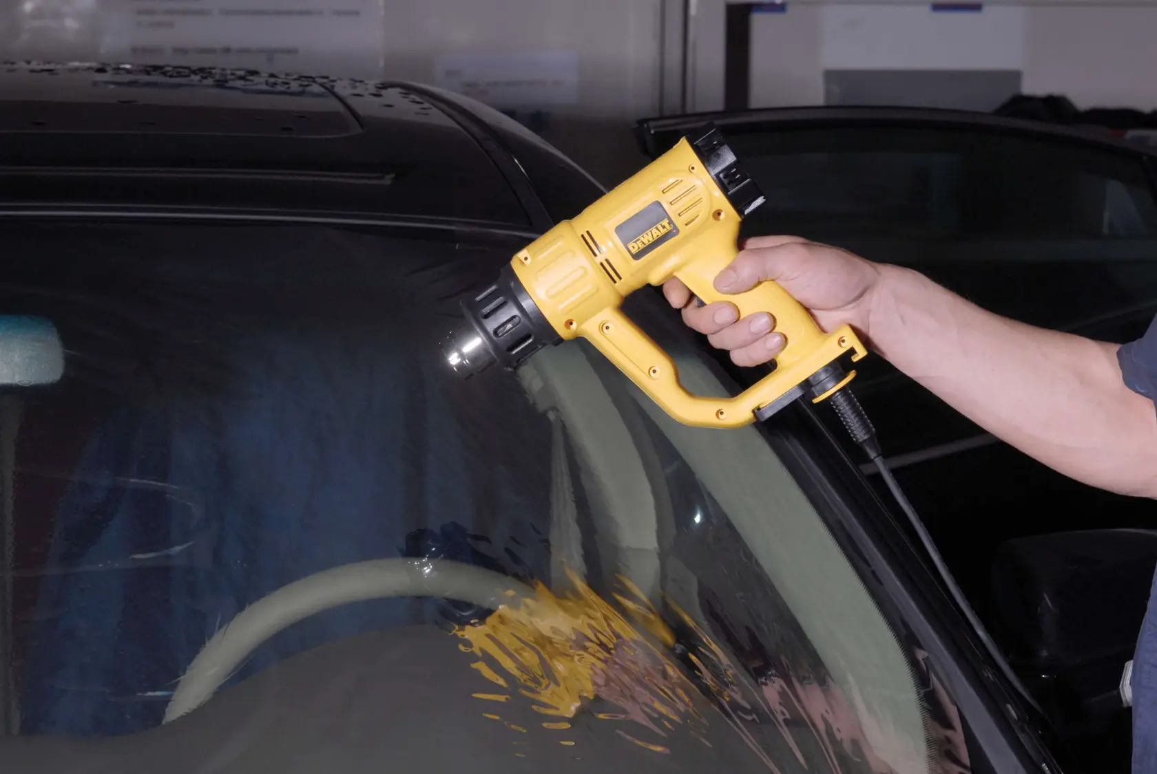Uma pessoa usando um soprador térmico DEWALT (modelo D26411) para aplicar calor no para-brisa de um carro.