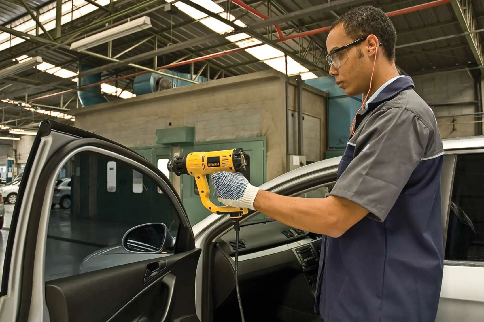 Uma pessoa usando luvas está utilizando um soprador térmico amarelo DEWALT (modelo D26411) no interior da porta de um carro em uma oficina. O rosto da pessoa está desfocado para preservar a privacidade.