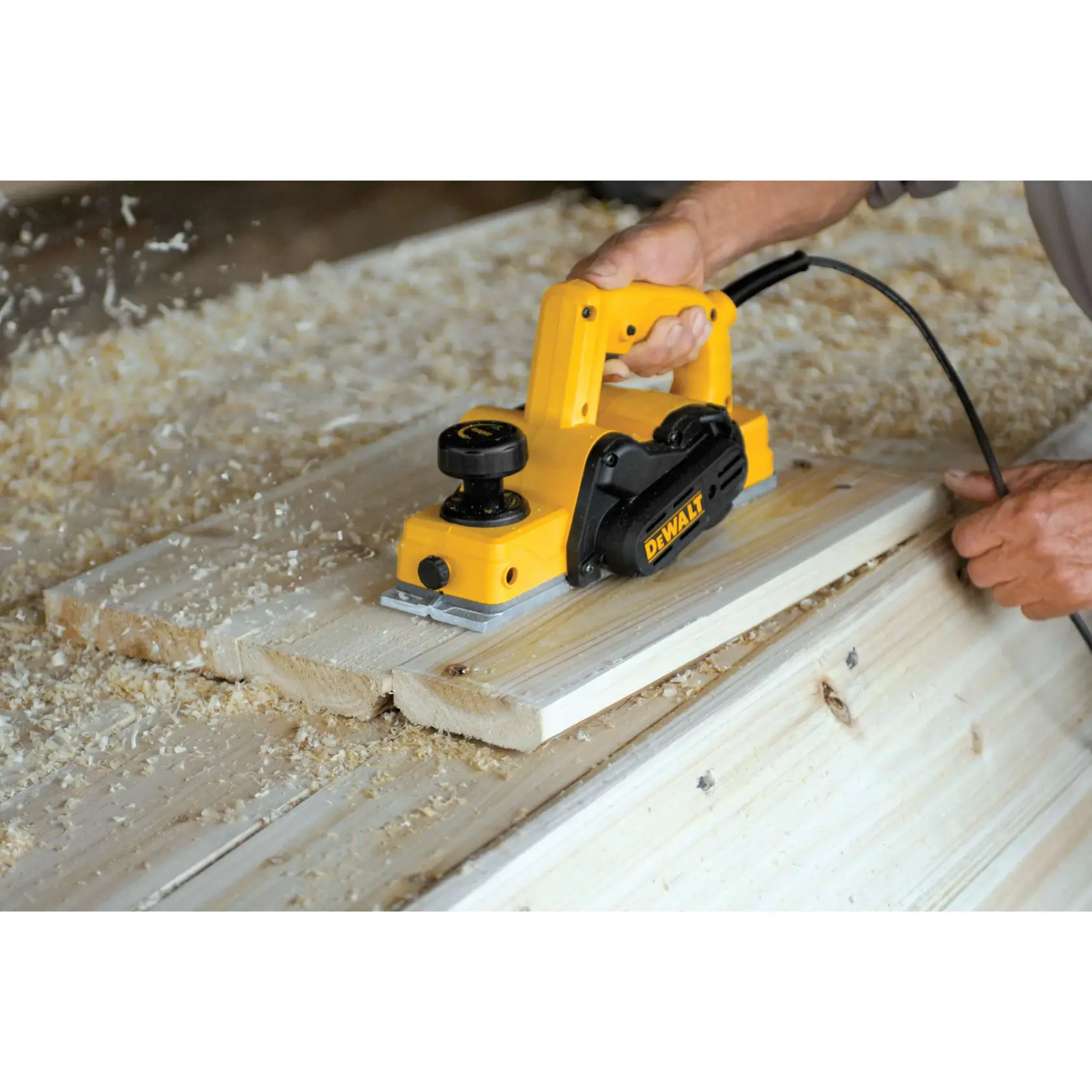 Uma pessoa utiliza uma plaina elétrica amarela DEWALT para nivelar tábuas de madeira, com lascas de madeira espalhadas pela área de trabalho.