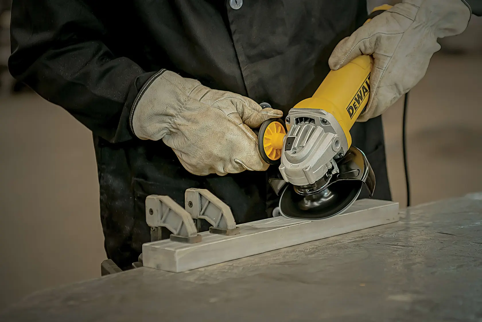 Pessoa usando luvas de proteção e casaco preto utilizando uma esmerilhadeira amarela DEWALT para trabalhar em uma peça de metal presa por grampos em uma bancada.