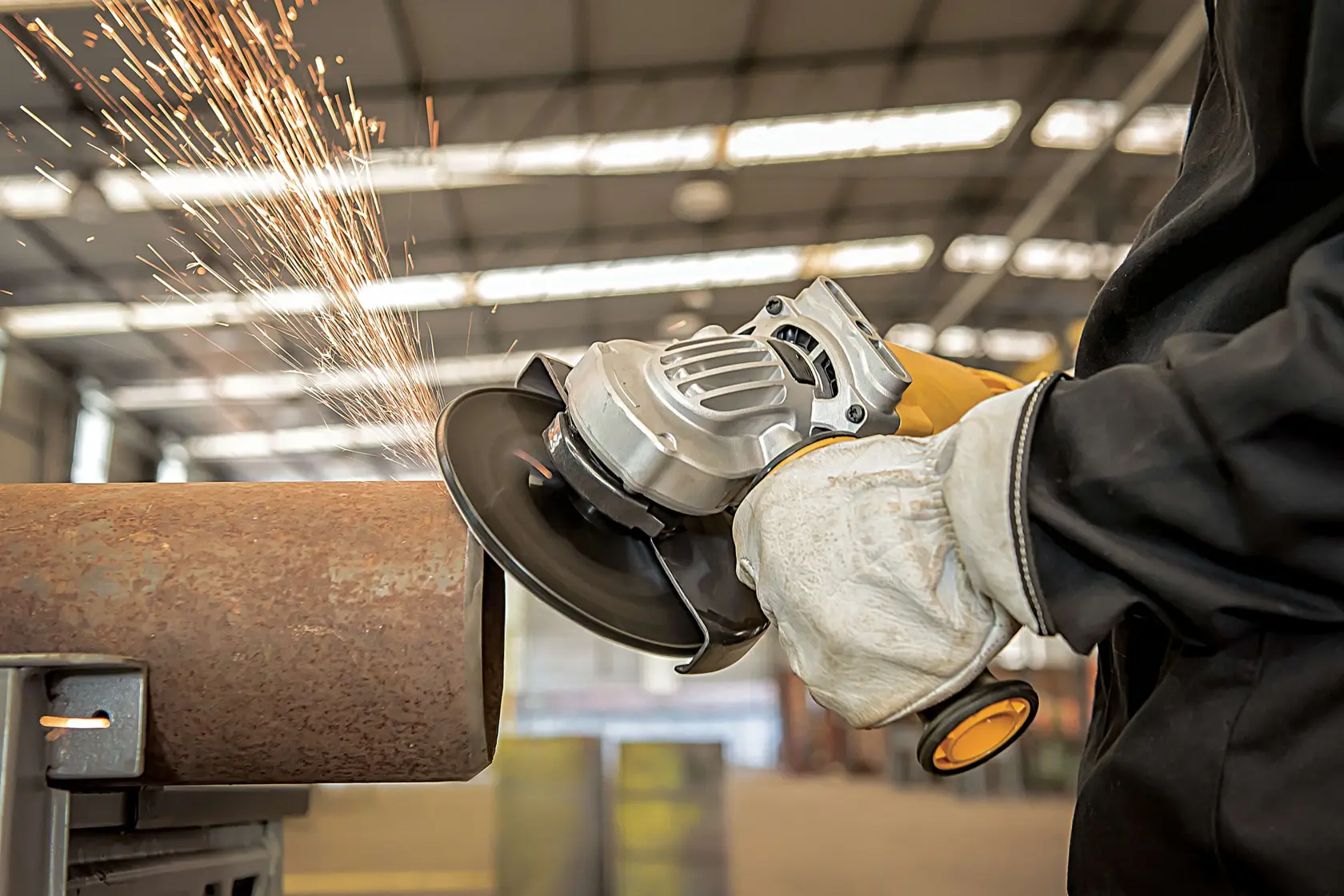 Uma pessoa usando luvas de proteção está utilizando uma esmerilhadeira angular DEWALT para cortar um tubo de metal, produzindo faíscas em um ambiente de oficina industrial.