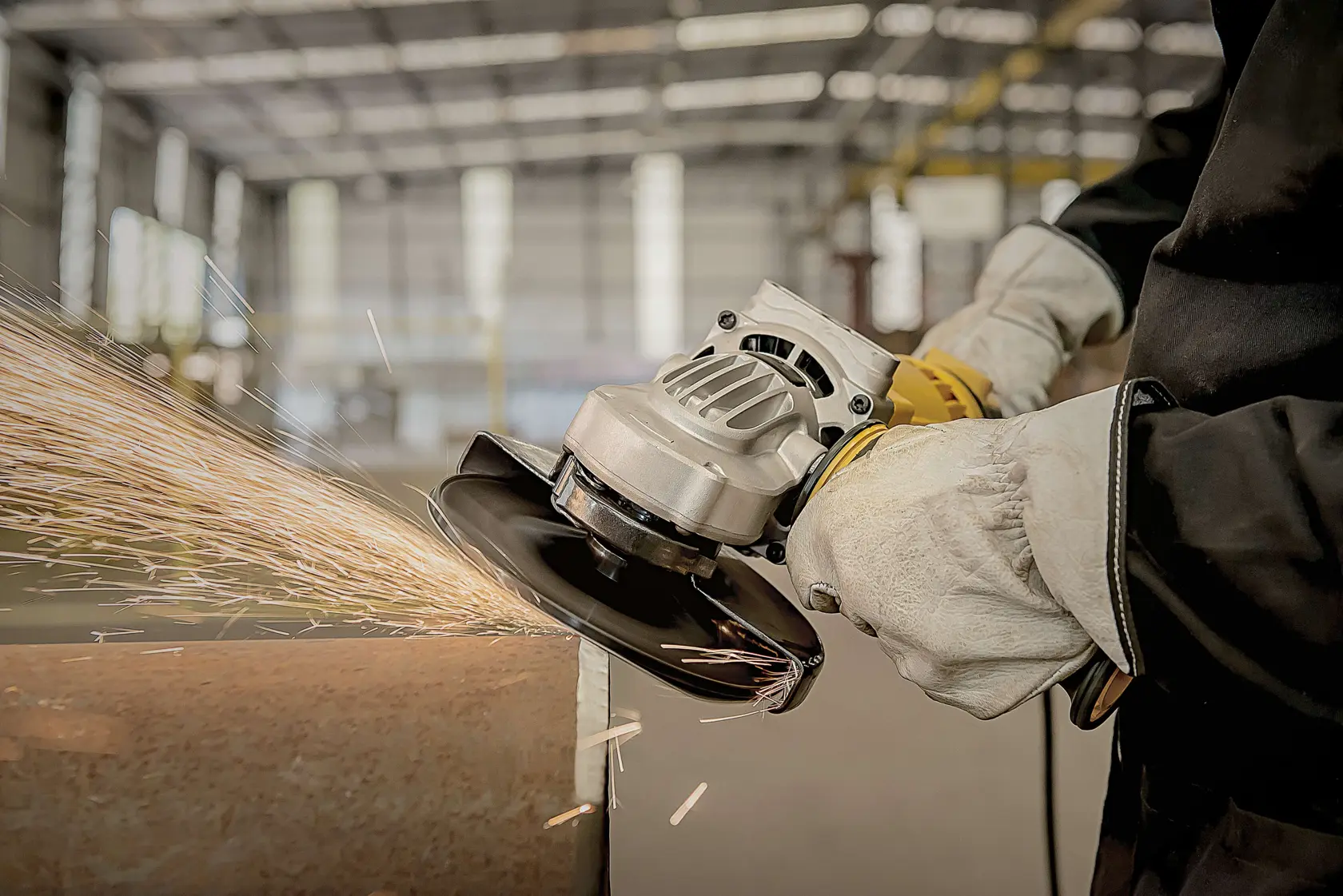 Close-up de uma pessoa usando uma esmerilhadeira angular DEWALT para cortar ou lixar uma superfície de metal, com faíscas voando. O usuário veste luvas de proteção e uma jaqueta de trabalho escura em um ambiente industrial.