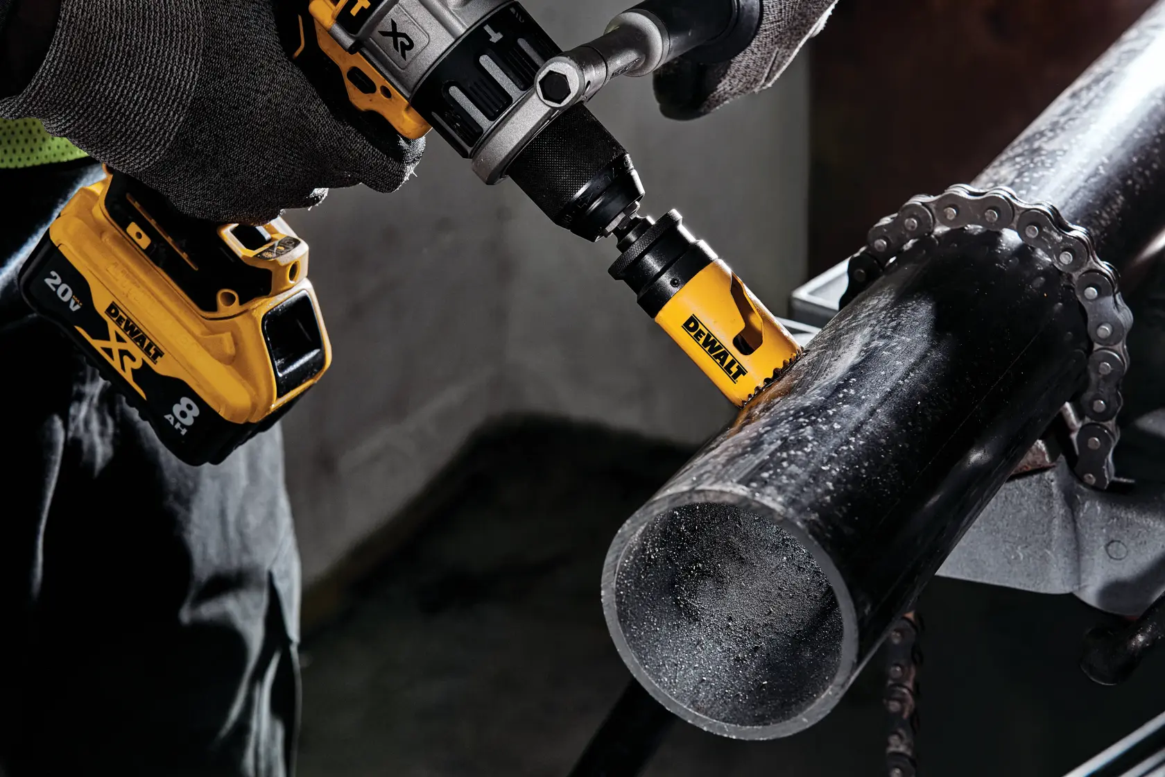 Imagem aproximada de uma pessoa usando uma furadeira sem fio DEWALT com acessório de serra copo para cortar um tubo de metal preto, com mãos enluvadas segurando a ferramenta firmemente.