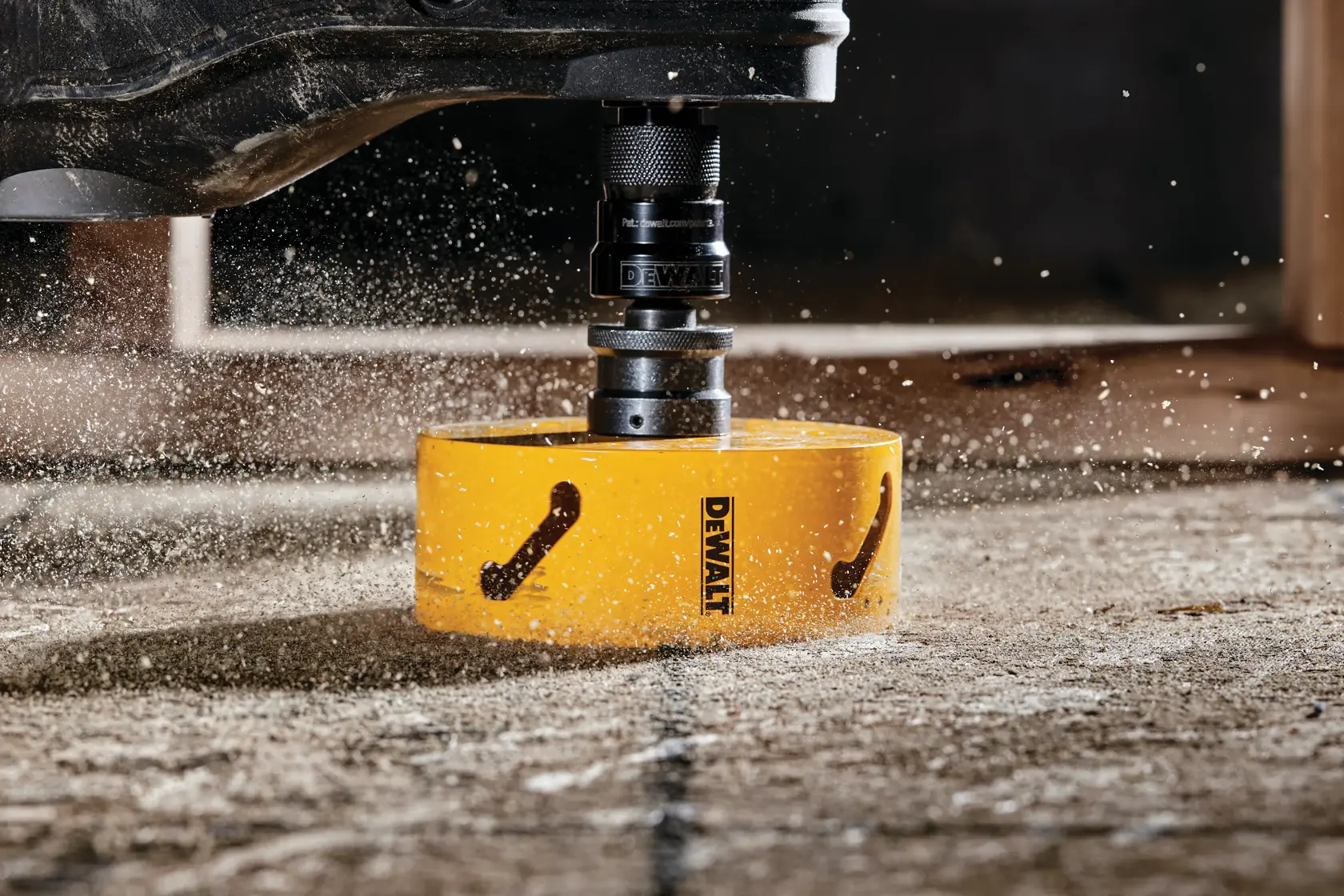 Close-up de um acessório de serra copo amarelo DEWALT cortando uma superfície de concreto, com poeira e detritos voando ao redor.