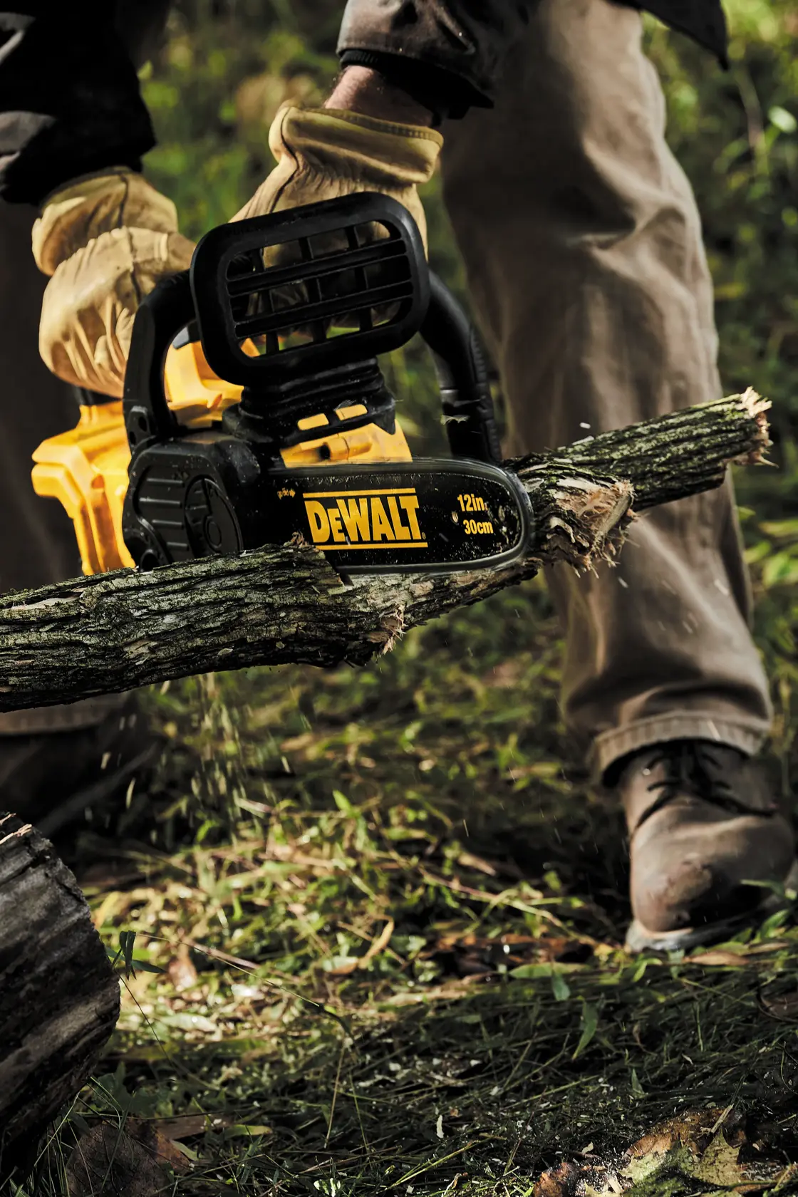 Uma pessoa usando luvas e roupas de trabalho está usando uma motosserra sem fio DEWALT para cortar um galho de árvore ao ar livre.