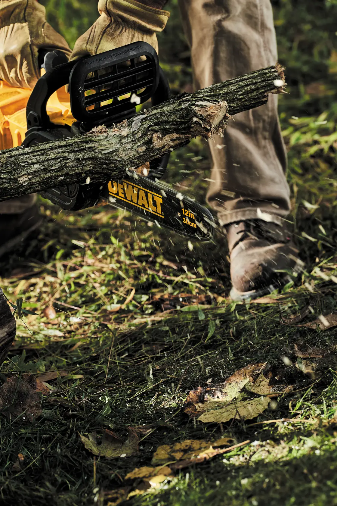 Uma pessoa usando luvas e roupas de proteção está utilizando uma motosserra DEWALT de 12 polegadas para cortar um galho de árvore ao ar livre, com lascas de madeira voando e grama embaixo.