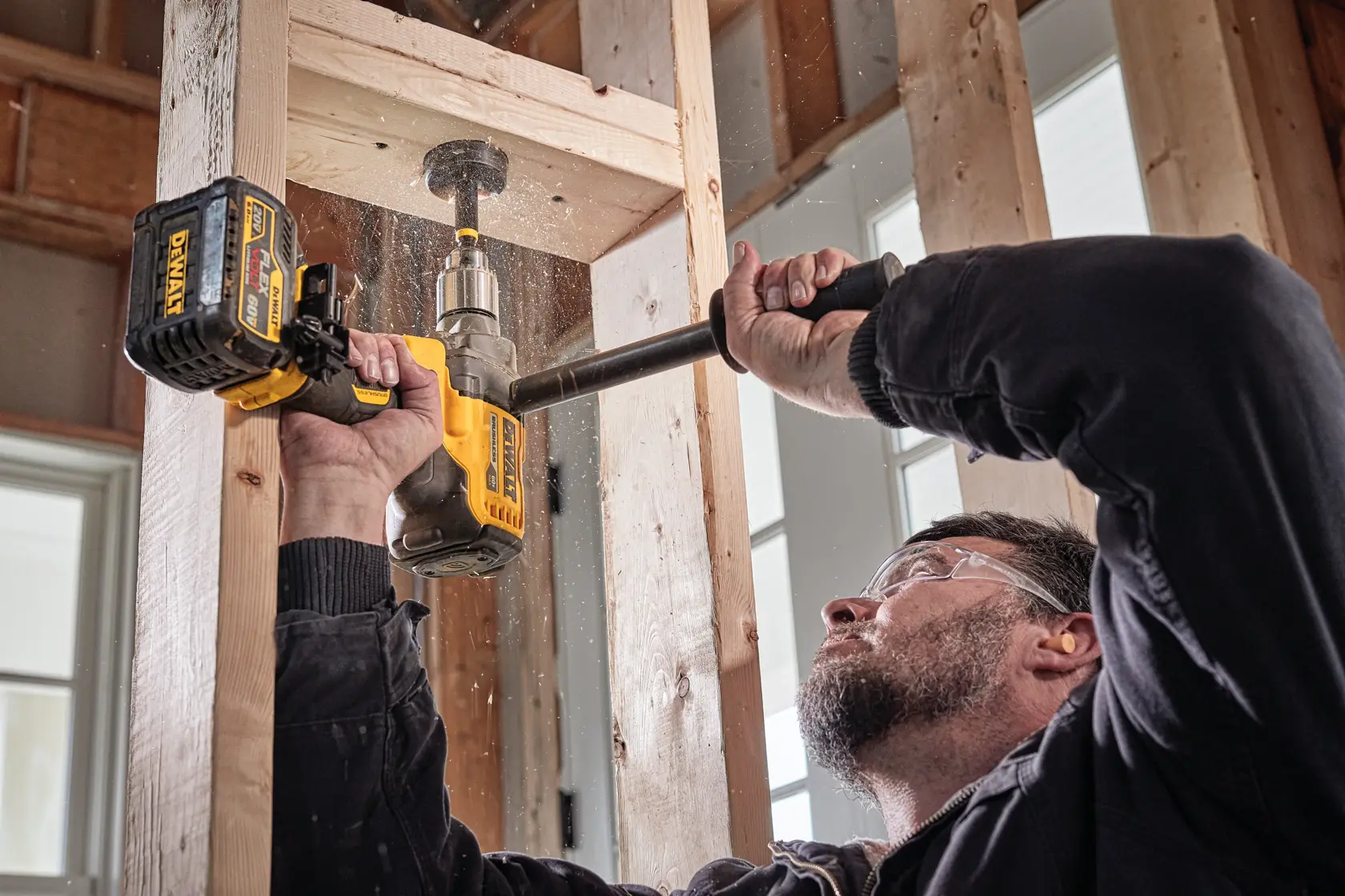 Pessoa usando uma furadeira sem fio DEWALT para fazer um furo em vigas de madeira dentro de um prédio em construção. A furadeira é segurada com as duas mãos e partículas de serragem são visíveis.