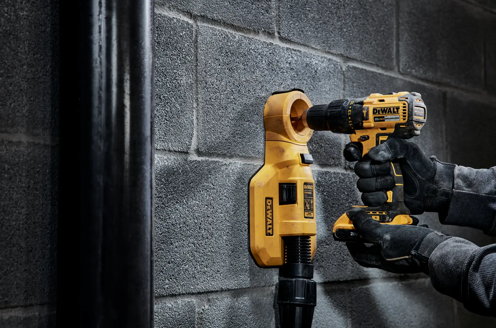 Uma pessoa usando luvas pretas está utilizando uma furadeira sem fio DEWALT para perfurar uma parede de blocos de concreto, com um acessório amarelo de extração de pó DEWALT conectado abaixo da furadeira.