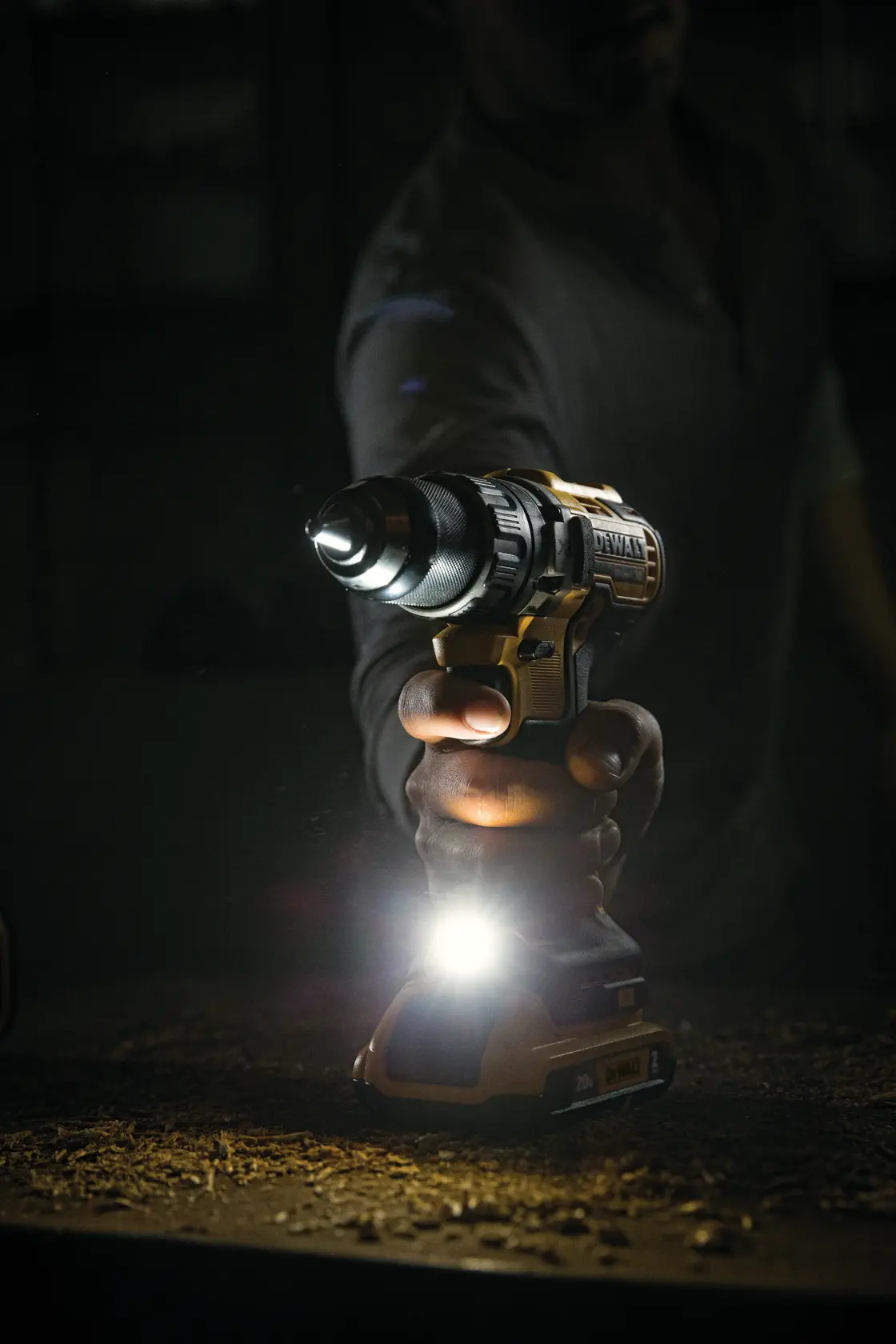 Uma pessoa segurando uma furadeira sem fio DEWALT com luz LED embutida acesa, iluminando a superfície de trabalho em um ambiente com pouca luz.
