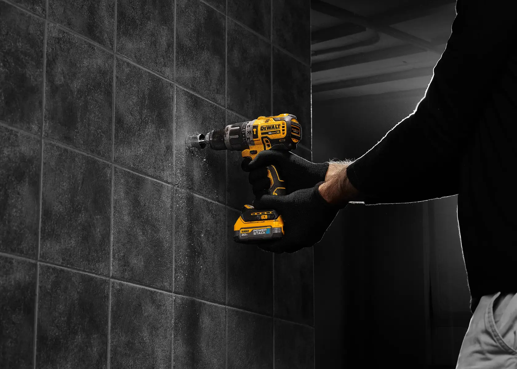 Uma pessoa usando luvas pretas utiliza uma furadeira sem fio amarela DEWALT para furar uma parede de azulejos escuros.