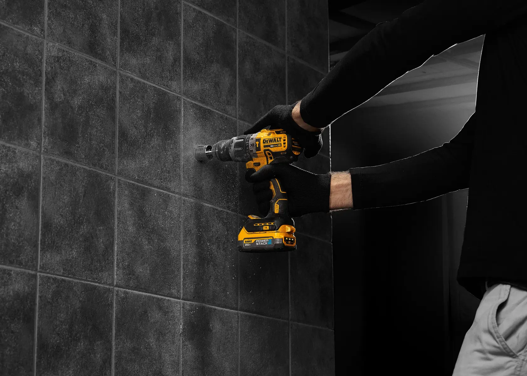 Uma pessoa com luvas pretas usa uma furadeira sem fio DEWALT para fazer um furo em uma parede de azulejos escuros.