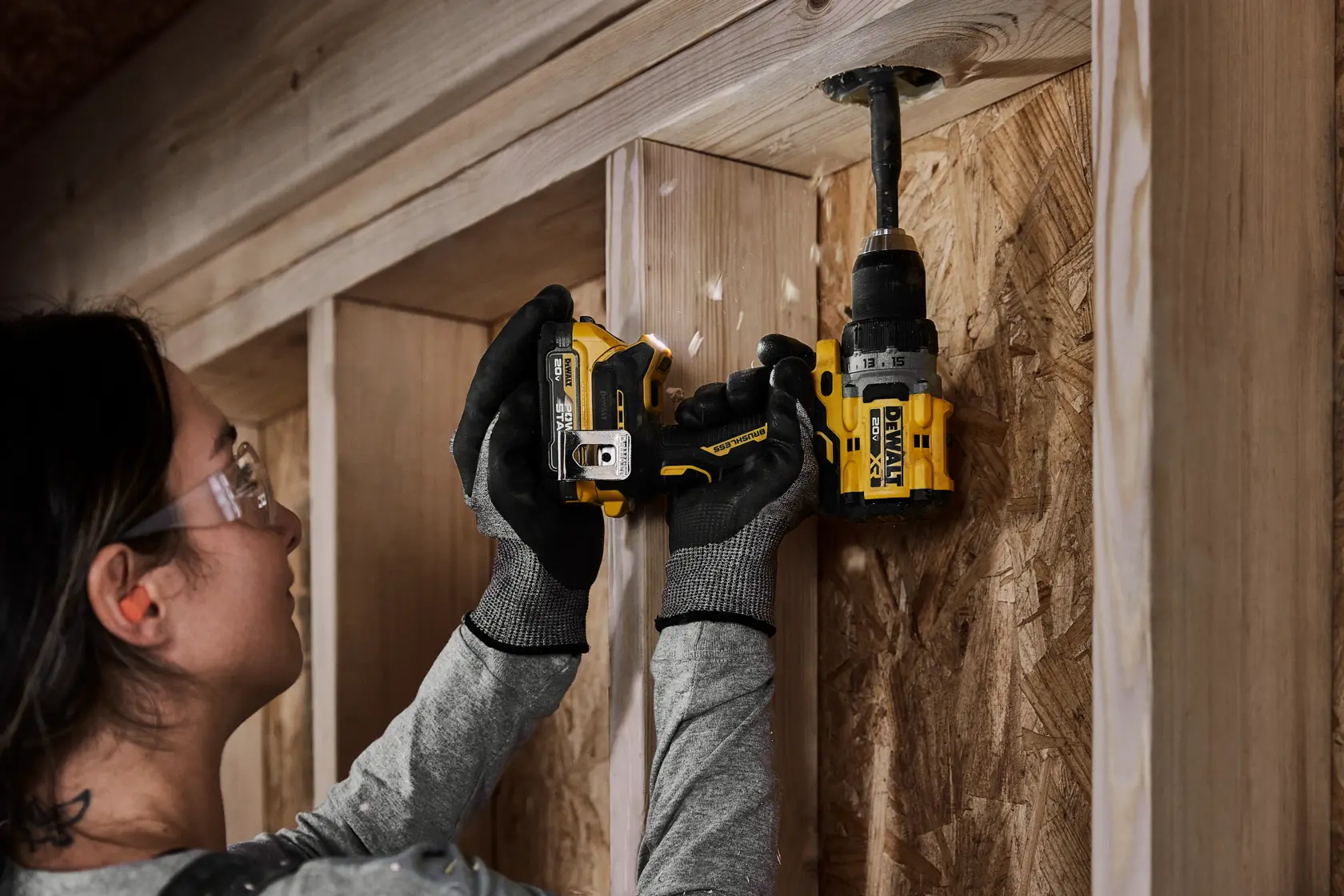 Uma pessoa usando luvas de trabalho está utilizando uma furadeira sem fio DEWALT para fazer um furo em uma estrutura ou parede de madeira.