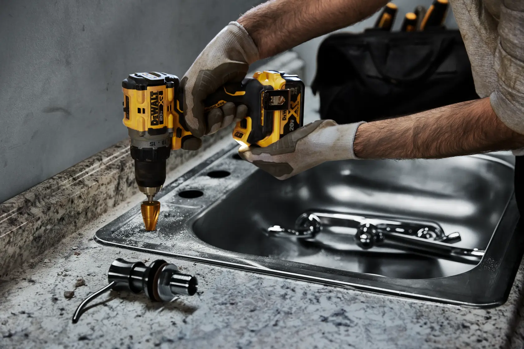 Uma pessoa usando luvas utiliza uma furadeira sem fio amarela e preta DEWALT para perfurar furos em uma bancada próxima a uma pia de aço inoxidável. Uma torneira e uma peça de encanamento são visíveis no balcão. Uma bolsa de ferramentas preta está ao fundo.