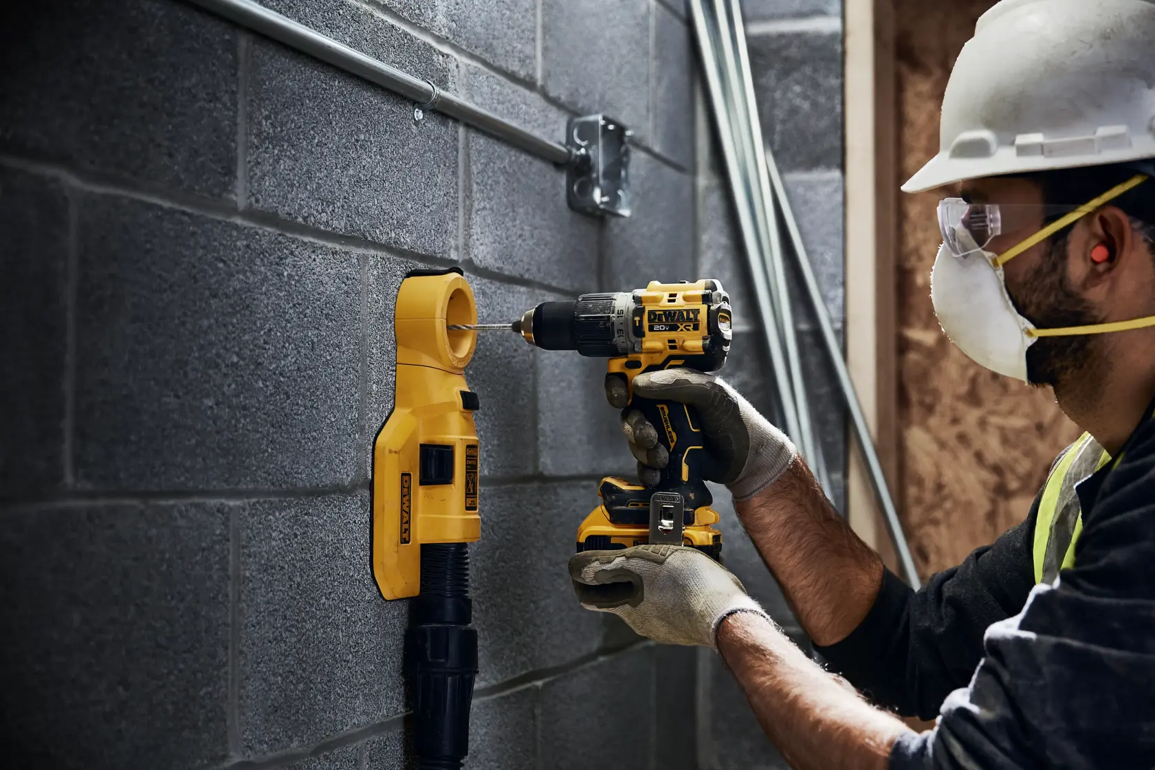 Uma pessoa usando equipamento de segurança utiliza uma furadeira sem fio DEWALT DCD805B em uma parede de concreto, com um acessório de extração de poeira DEWALT montado abaixo do local de perfuração.