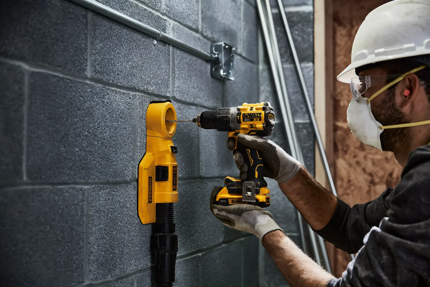 Uma pessoa usando luvas de trabalho e capacete está utilizando uma furadeira sem fio amarela e preta DEWALT para perfurar uma parede de blocos de concreto. Um acessório extrator de pó amarelo DEWALT está posicionado na parede abaixo da furadeira.