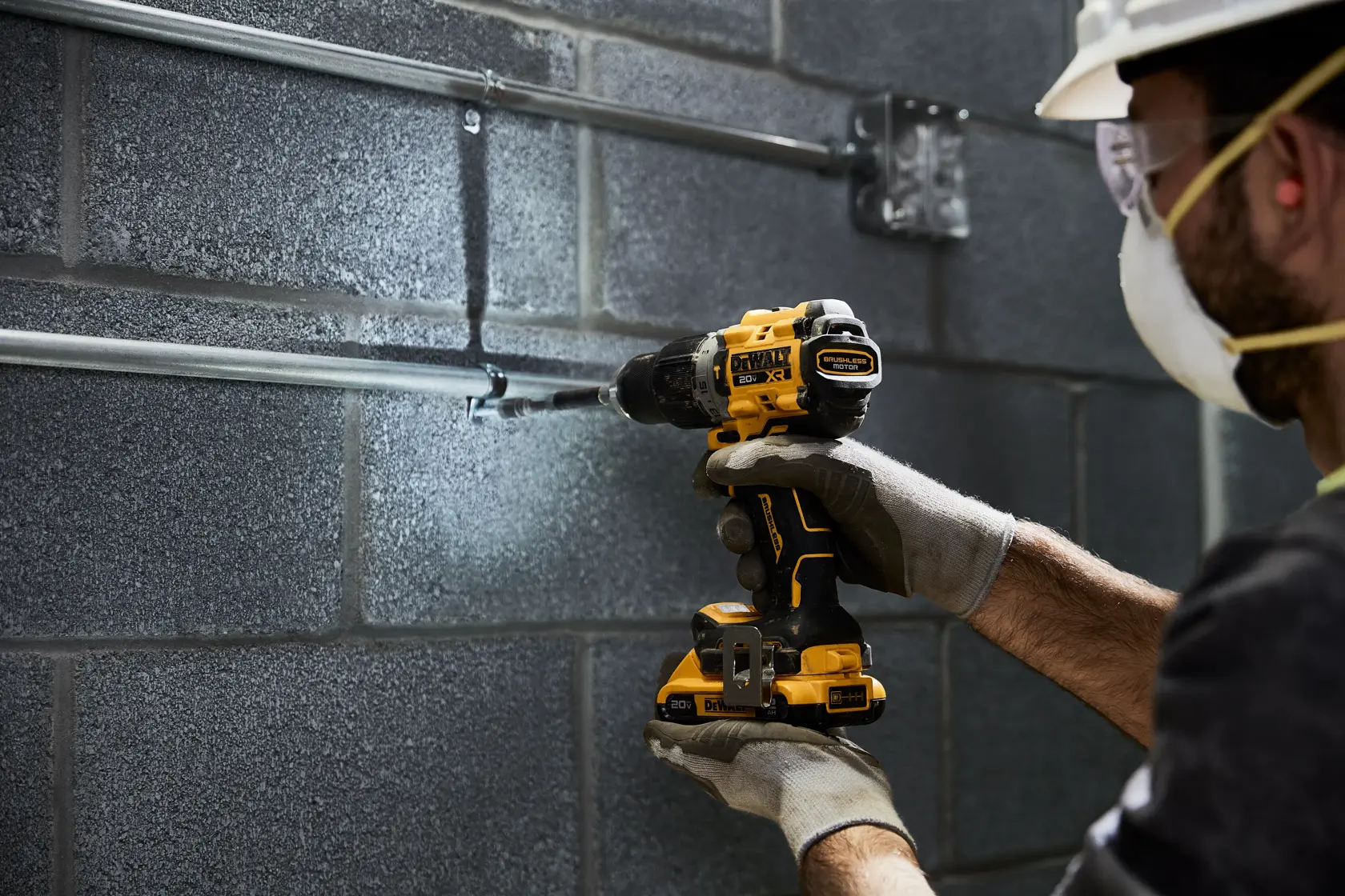 Uma pessoa usando luvas e capacete de segurança utiliza uma furadeira sem fio amarela e preta DEWALT para trabalhar em tubos metálicos fixados a uma parede de blocos cinza.