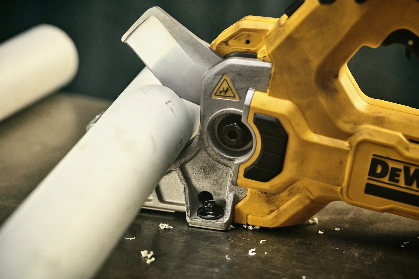 Uma pessoa usando luvas de trabalho está utilizando um cortador de tubos sem fio amarelo DEWALT para cortar um tubo de PVC branco, com texto indicando que a ferramenta pode cortar PVC Schedule 80 de 2 polegadas em até 3 segundos. A bateria da ferramenta é mostrada e observada como vendida separadamente.
