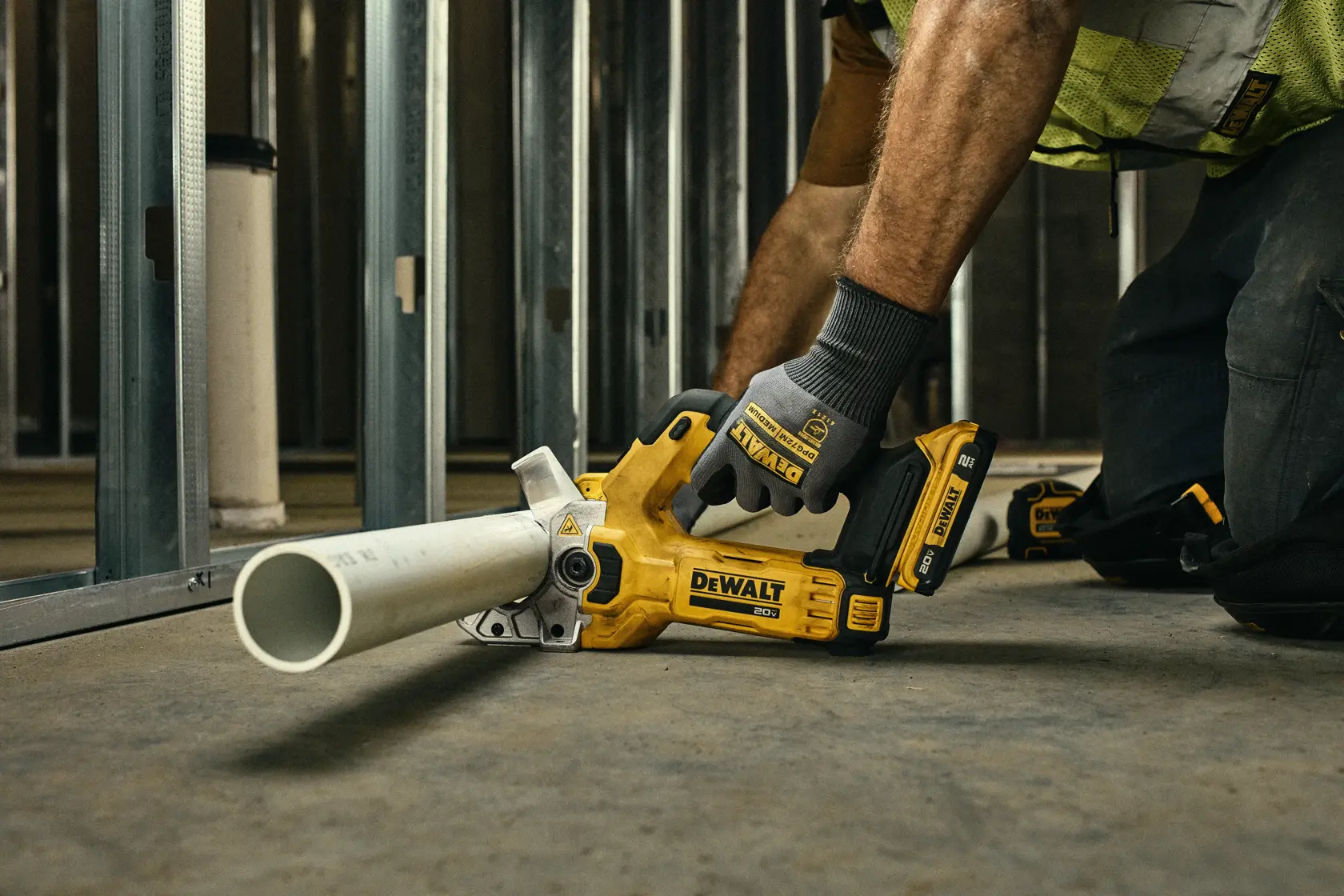 Uma pessoa usando luvas de trabalho está cortando um tubo plástico branco com um cortador de tubos amarelo sem fio da DEWALT em um ambiente industrial com uma parede de blocos de concreto e caixas de ferramentas.