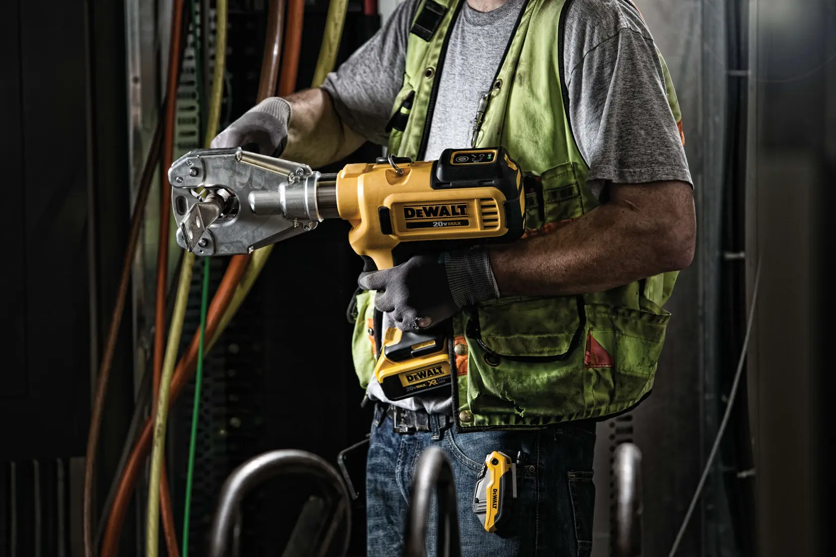 Uma pessoa usando um colete de segurança verde e luvas está utilizando uma ferramenta de crimpagem sem fio DEWALT, modelo DCE350M2, em um ambiente industrial com cabos e equipamentos ao fundo.