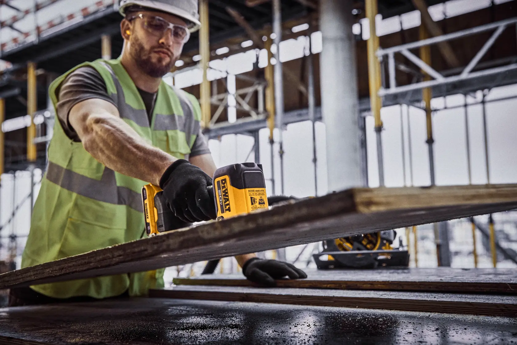 Um trabalhador da construção civil usando colete de segurança e capacete está carregando uma grande folha de madeira compensada fora de um prédio em construção. O trabalhador está usando um cinto de ferramentas e uma ferramenta elétrica amarela DEWALT presa na cintura.