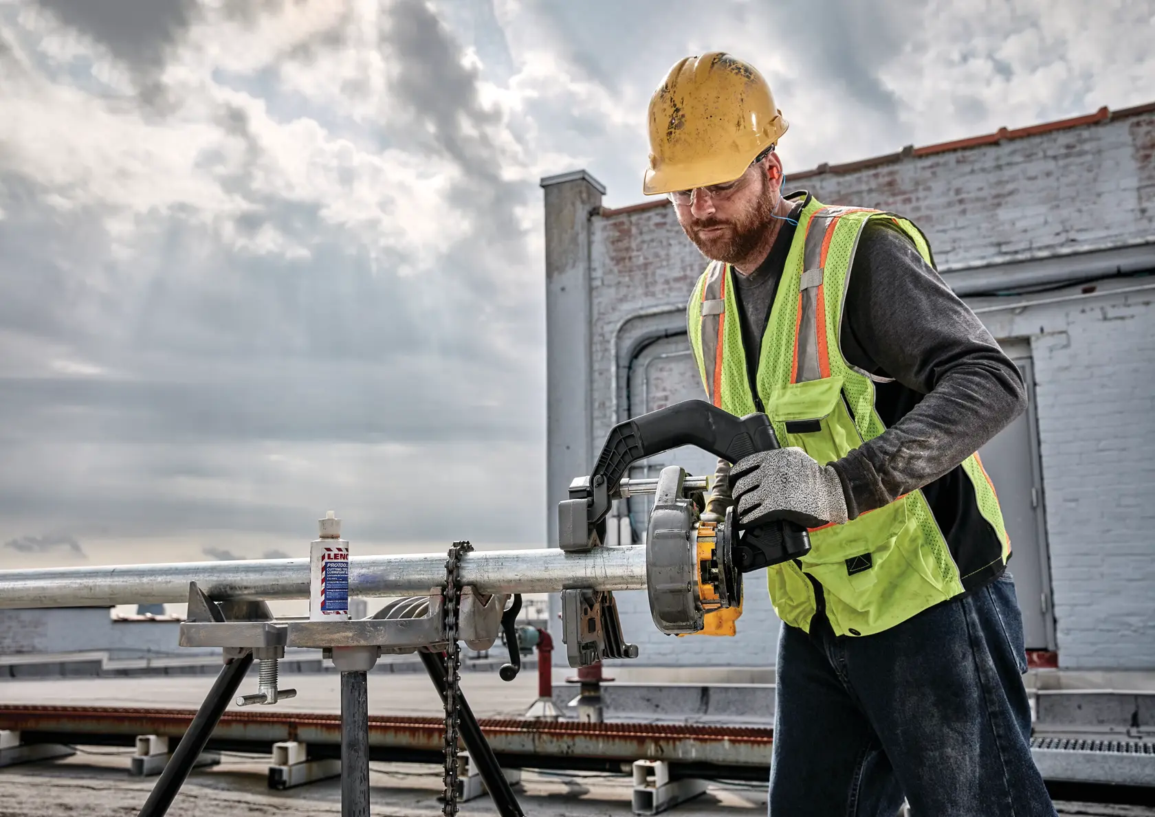 Um trabalhador da construção civil usando capacete amarelo, colete de segurança e luvas opera uma ferramenta de rosca DEWALT em um tubo de metal em um canteiro de obras ao ar livre. O trabalhador está usando a ferramenta para roscar o tubo, que está fixado em um suporte, com céu nublado e ambiente industrial ao fundo.