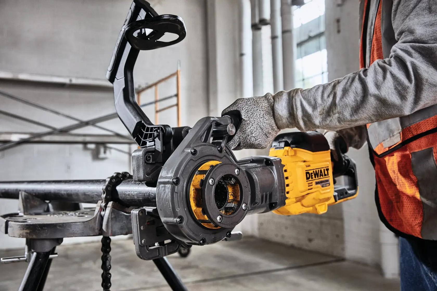 Uma pessoa usando luvas de segurança e colete opera uma ferramenta de roscagem de tubos DEWALT em um tubo de metal em um ambiente industrial.