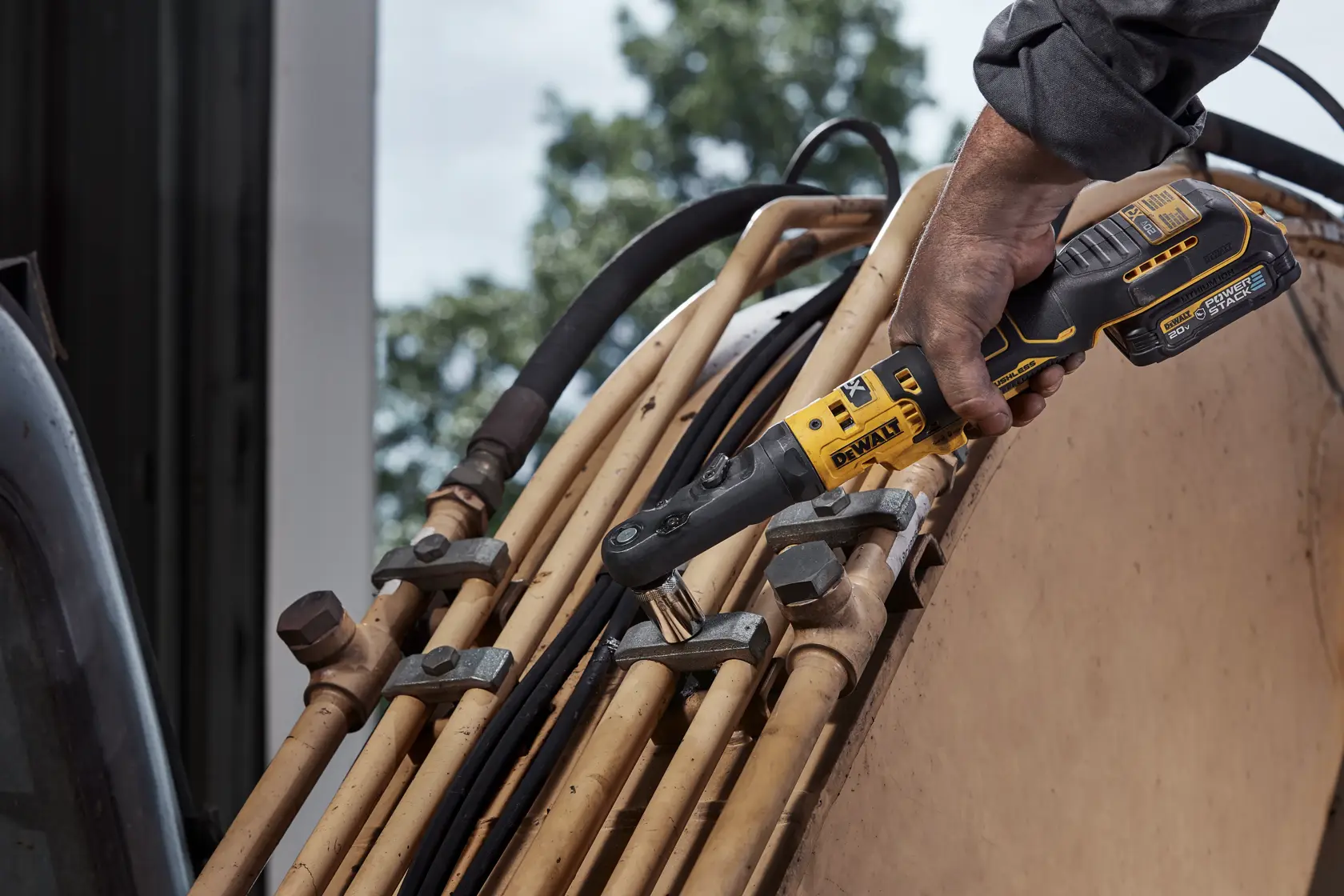 Close-up de mãos sujas segurando uma ferramenta elétrica DEWALT com bigornas intercambiáveis, em uma bancada de oficina com parafusos e peças mecânicas espalhadas. O texto 'INTERCHANGEABLE ANVILS' aparece em letras amarelas na parte inferior.