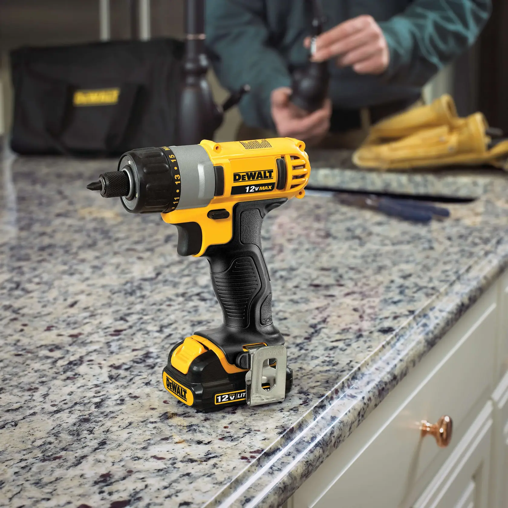 Uma furadeira sem fio DEWALT 12V MAX com bateria, em pé sobre uma bancada de granito em uma cozinha. Ao fundo, uma pessoa está trabalhando com algumas ferramentas e luvas.