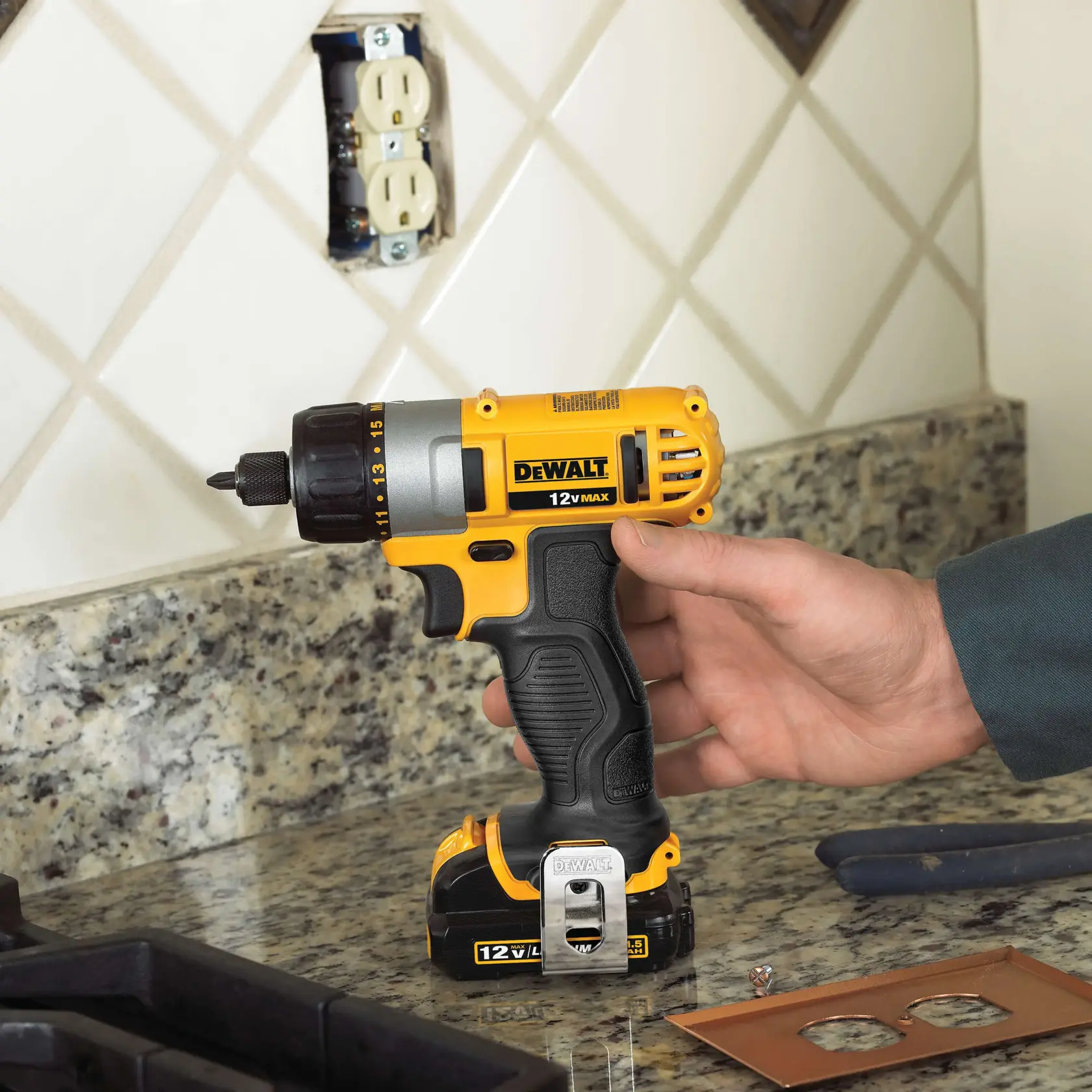 Uma pessoa segura uma furadeira sem fio DeWalt 12V MAX amarela e preta sobre uma bancada de granito na cozinha, com um backsplash de azulejos. A furadeira está perto de uma tomada elétrica sendo trabalhada e algumas ferramentas e tampas de tomadas estão próximas.