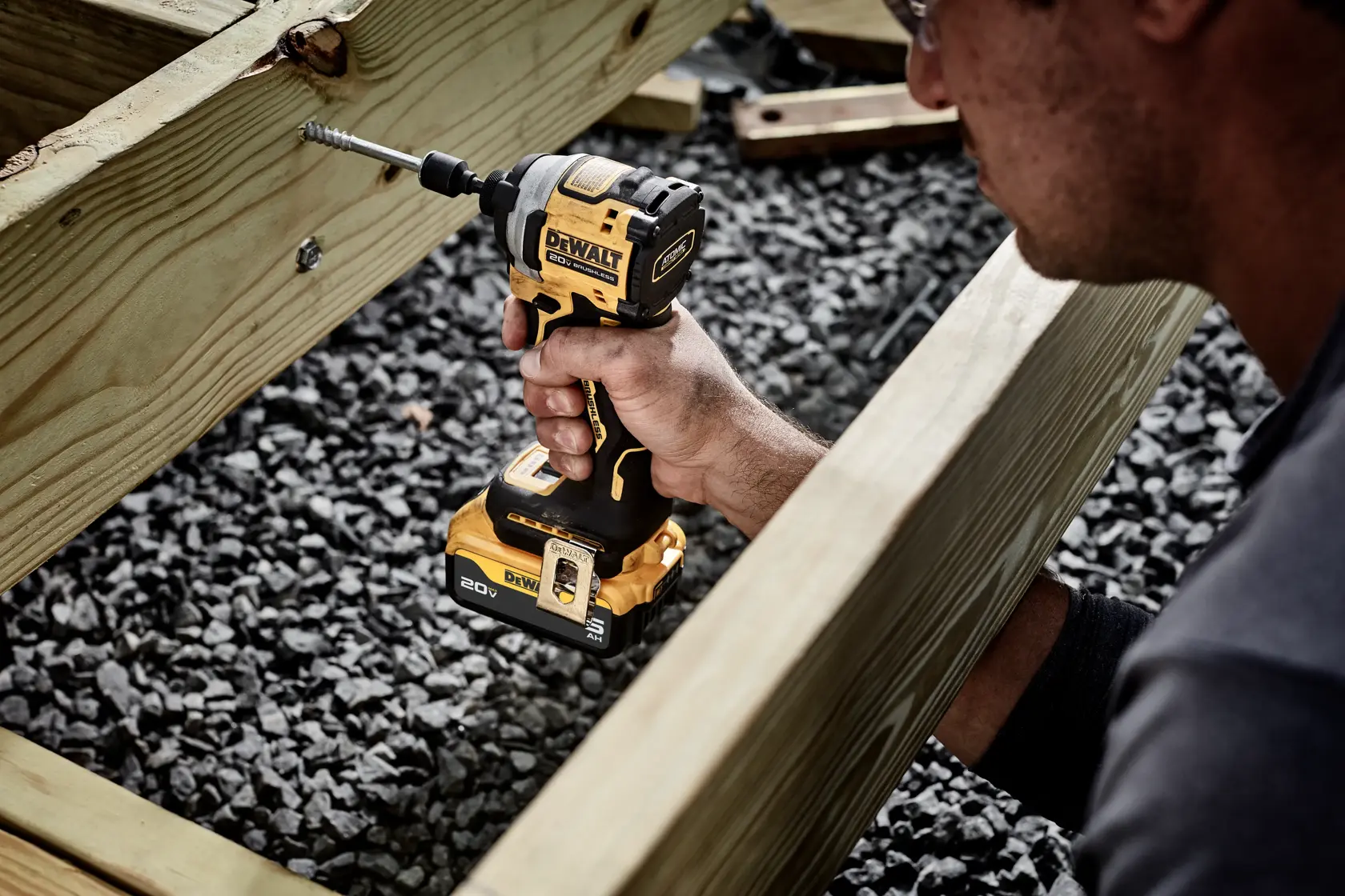 Uma pessoa utiliza um parafusador de impacto sem fio DEWALT DCF850B para parafusar em uma estrutura de madeira, com cascalho embaixo.