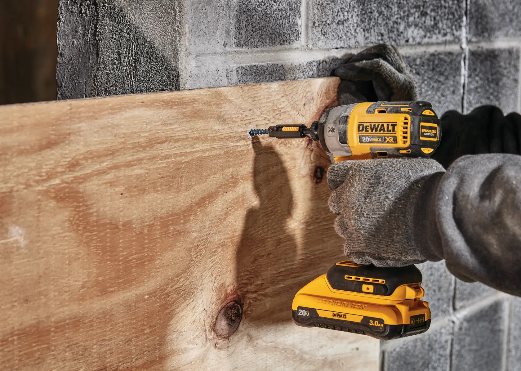 Uma pessoa usando luvas utiliza uma parafusadeira de impacto sem fio DEWALT 20V Max XR amarela e preta para parafusar um parafuso em uma tábua de madeira presa a uma parede de concreto.