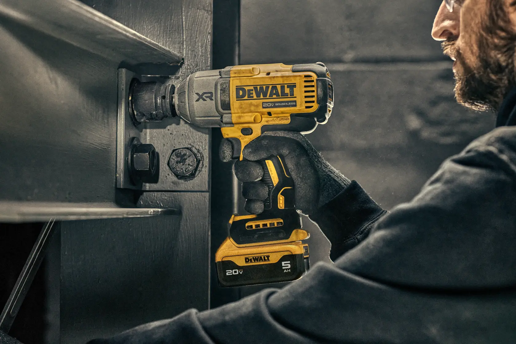 Uma pessoa usando luvas utiliza uma chave de impacto sem fio DEWALT de 20V para apertar um parafuso em uma grande estrutura metálica.