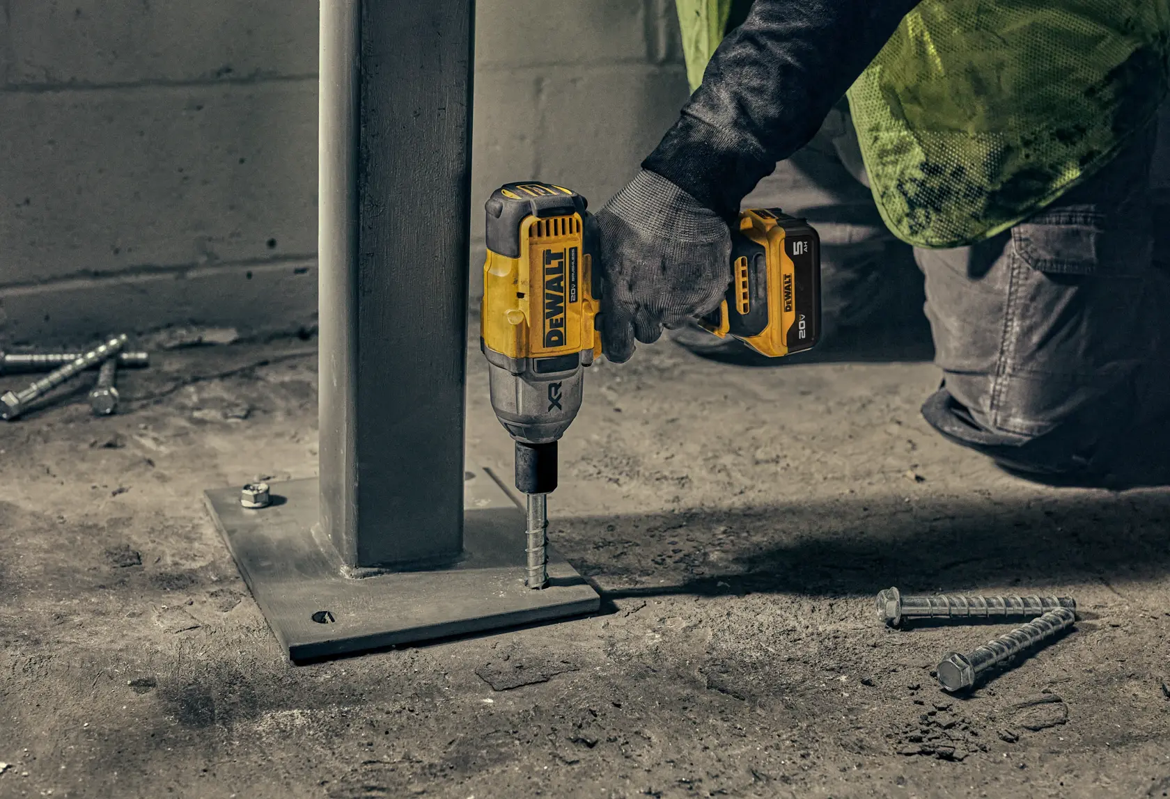 Uma pessoa utiliza uma parafusadeira de impacto sem fio DEWALT para fixar um parafuso na base de uma coluna de metal. Vários parafusos estão visíveis no chão ao redor, e a pessoa está usando luvas e calça de trabalho.