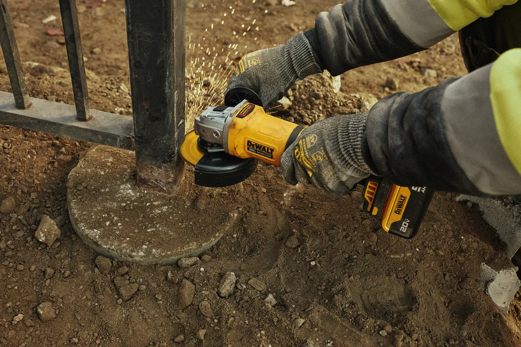 Uma pessoa usando luvas usa uma esmerilhadeira angular sem fio amarela DEWALT para cortar vergalhões de aço que saem de um bloco de concreto, com faíscas visíveis durante o processo de corte.