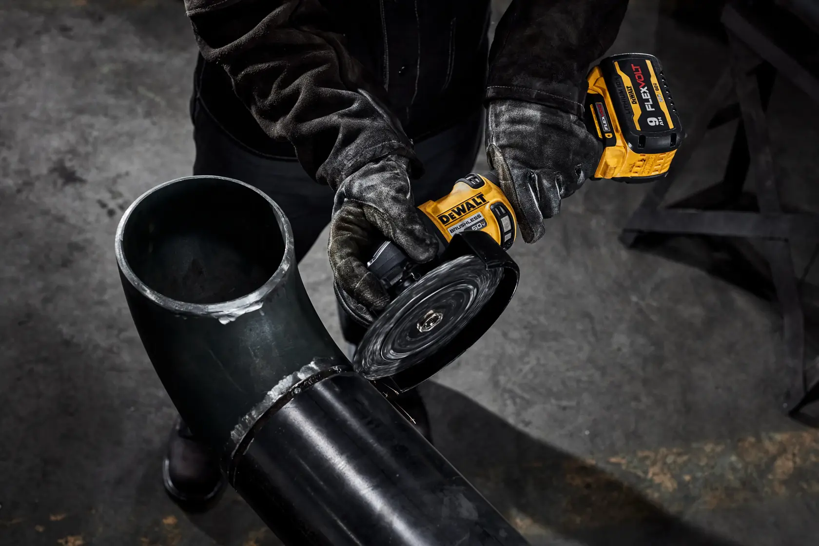 Uma pessoa usando luvas de proteção está utilizando uma esmerilhadeira sem fio DEWALT para cortar ou polir um grande tubo de metal preto em um ambiente industrial.
