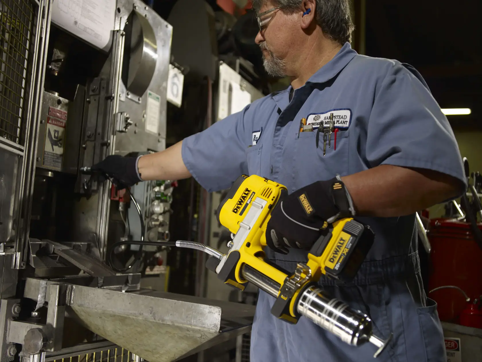 Um trabalhador opera uma máquina industrial segurando uma pistola de graxa sem fio DEWALT DCGG571M1, usando luvas de segurança e uniforme azul.