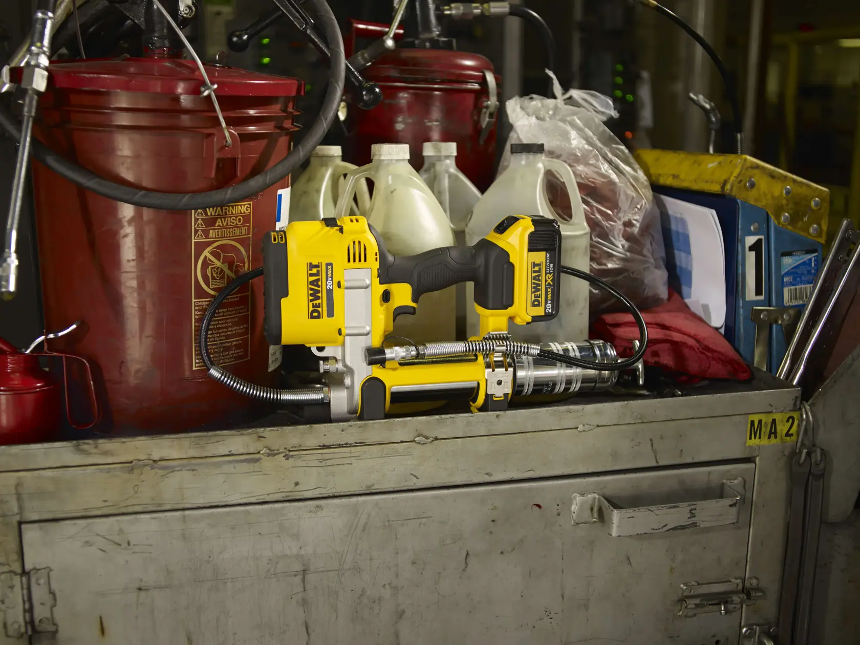 Uma pistola de graxa sem fio DEWALT modelo DCGG571M1 está colocada sobre um gabinete de oficina de metal, cercada por grandes recipientes vermelhos, garrafas plásticas e várias ferramentas e suprimentos.