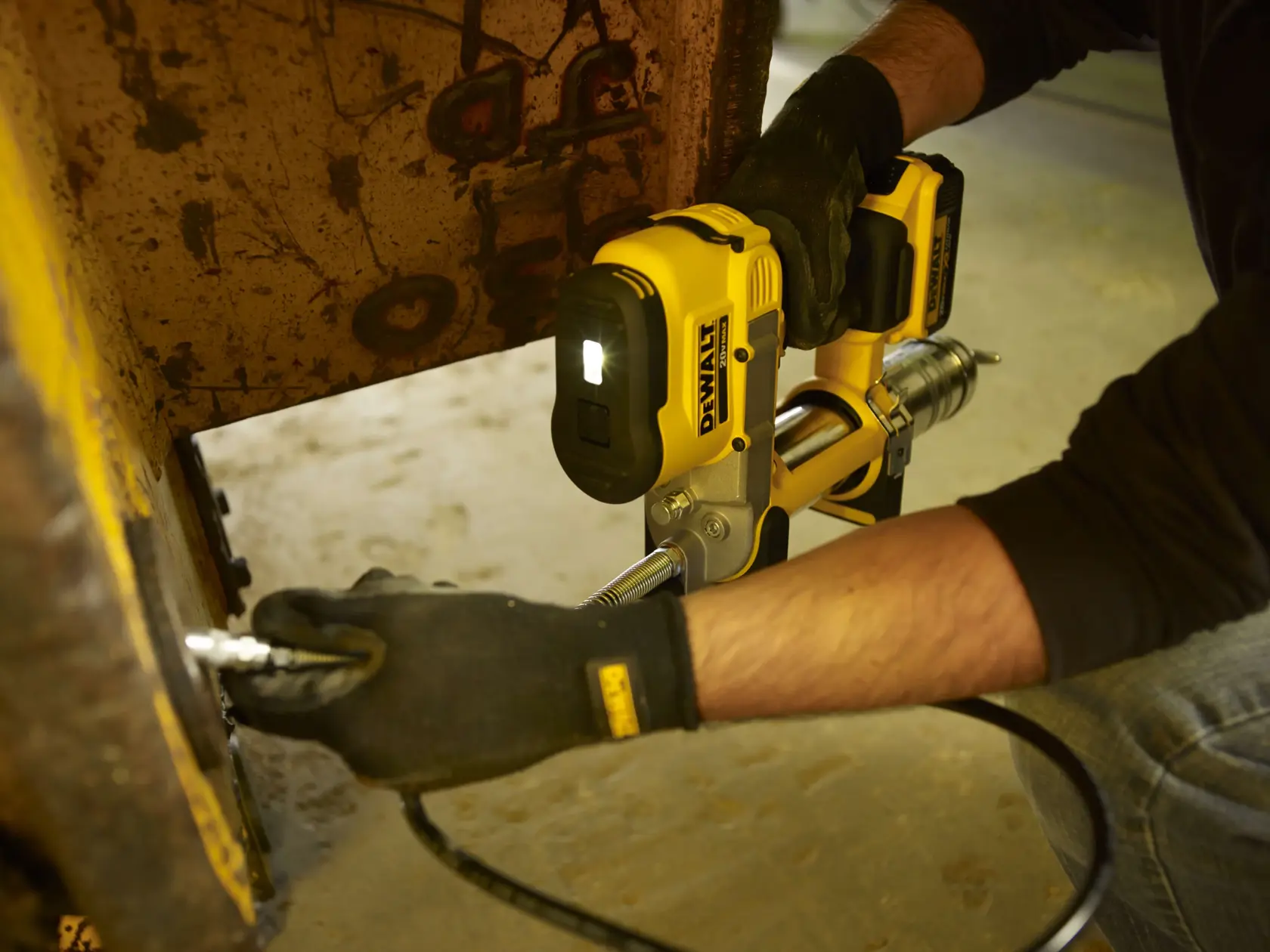 Uma pessoa usando luvas utiliza uma pistola de graxa sem fio DEWALT para lubrificar uma máquina, com a luz LED da ferramenta ligada para melhor visibilidade.